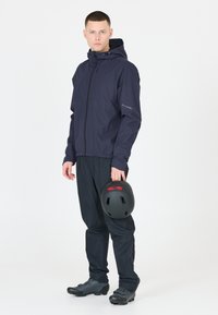 Marinblå vattentät jacka med huva, framdragkedja och reflexdetaljer. Svart byxor och skor. Håller en svart hjälm med röd accent.