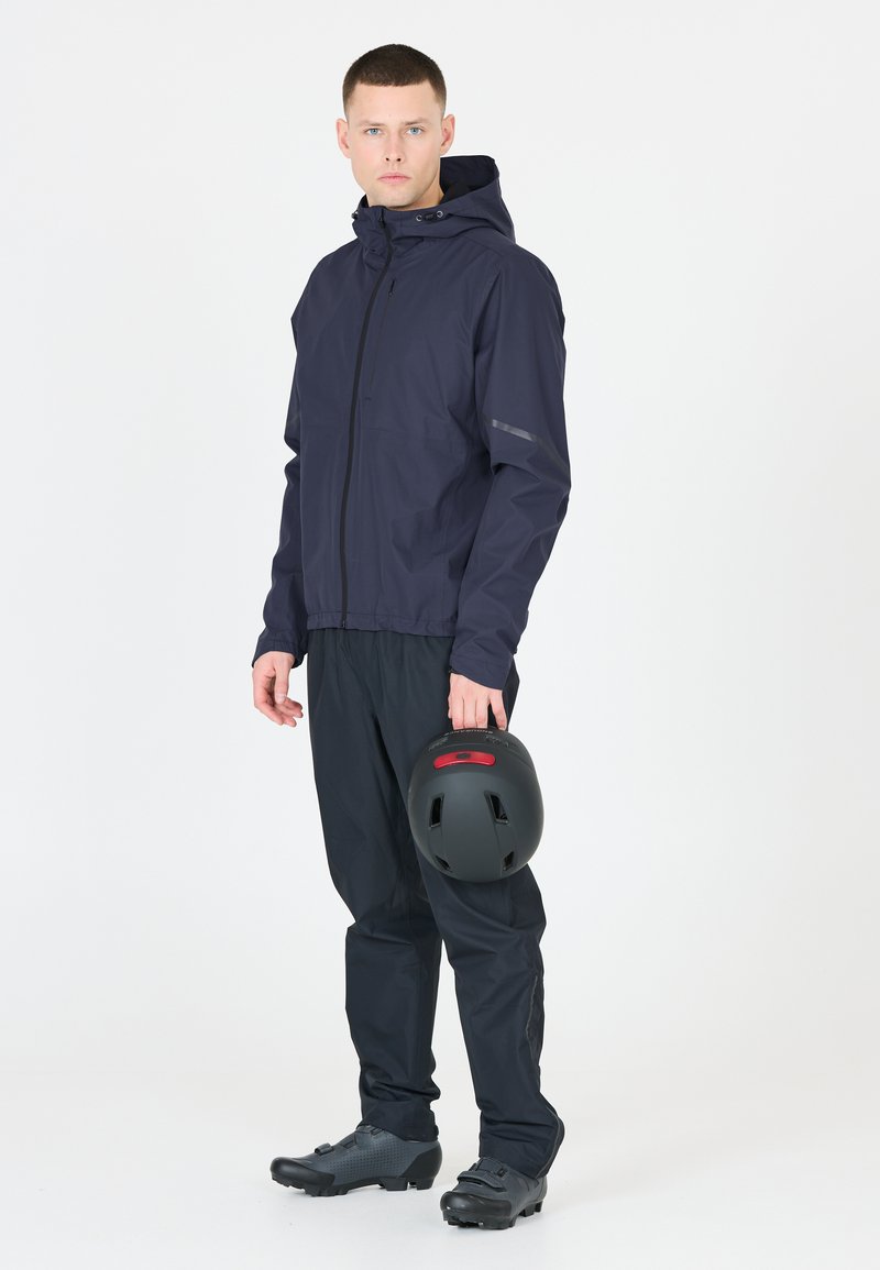 Marinblå vattentät jacka med huva, framdragkedja och reflexdetaljer. Svart byxor och skor. Håller en svart hjälm med röd accent.