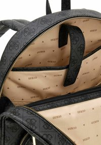 Sac à dos noir en synthétique avec fermeture éclair, présentant un extérieur texturé et un intérieur beige à motif à pois, ainsi que plusieurs compartiments.