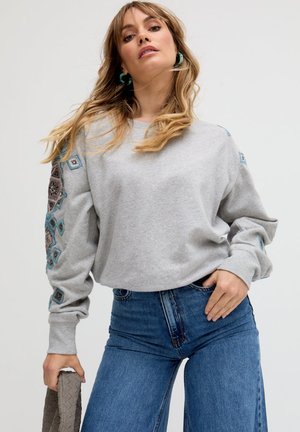 Frau trägt ein hellgraues Sweatshirt mit blau-lila gemusterten Stickereien an den Ärmeln, kombiniert mit hoch taillierten blauen Jeans, hält eine strukturierte Clutch.