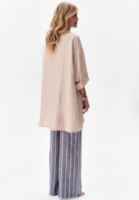 Beżowa, oversize koszula z podwiniętymi rękawami, zestawiona z niebieskimi, szerokimi spodniami w paski z żółtymi liniami. Miękki materiał o casualowym stylu.