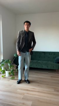 Braunes doppeltknopfliges Samtblazer über einem schwarzen Hemd, hellblauen Jeans und schwarzen Schuhen. Grünes Sofa und Topfpflanze im Hintergrund.