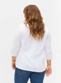 Weißes, langärmliges T-Shirt mit lockerer Passform und leicht gebogenem Saum. Kombiniert mit blauen Denim-Jeans, von hinten betrachtet.