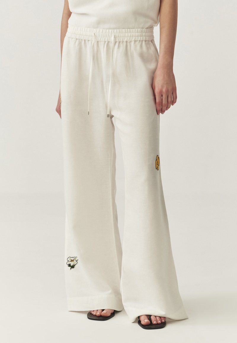 Pantalons larges en lin blanc avec une taille à cordon, ornés de motifs brodés jaunes et verts sur les jambes. Texture douce et coupe décontractée.