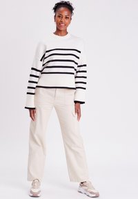 Pull en maille épaisse blanc avec des rayures horizontales noires, associé à un pantalon large beige et des baskets légères. Texture douce et coupe décontractée.
