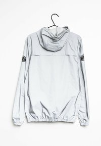 Ljusgrå vindjacka med huva, elastiska ärmslut och nederkant, samt svarta logopatchar på båda ärmarna, hängande på en svart galge.