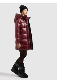 Mujer con abrigo acolchado burdeos con capucha, gorro negro, medias negras y botas de invierno negras, de pie frente a un fondo liso.