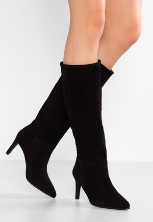 Kobieta nosząca czarne zamszowe kozaki do kolan z zaostrzonymi noskami i smukłymi wysokimi obcasami, stojąca na białej powierzchni.