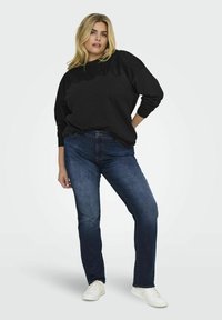Černá mikina s texturovaným hrdlem, spárovaná s tmavě modrými klasickými džíny. Model má na sobě bílé tenisky a stojí na neutrálním pozadí.