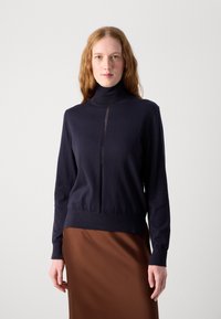 Marineblå turtleneck genser i et mykt stoff, med sømdetaljer foran og ribbestrikkede mansjetter og hem. Kombinert med et brunt skjørt.
