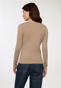 Maglione beige a maniche lunghe con collo rotondo, polsini e orlo a coste, realizzato in un tessuto a maglia morbido. Indossato con pantaloni in denim scuri.