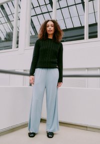 Donna con capelli ricci si trova all'interno, indossando un pullover nero a maglia cablata, pantaloni a gamba larga blu chiaro e scarpe nere, vicino a pareti e finestre bianche.