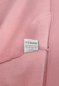 Giaccone in pile rosa chiaro con un'etichetta che mostra le istruzioni per la cura e il nome del marchio. Texture morbida, polsini e dettagli di cucitura visibili.