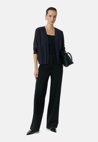 Czarne szerokie spodnie zestawione z ciemną, lekką bluzką zapinaną na otwartym przodzie. Modelka trzyma małą czarną torebkę, o gładkich teksturach.