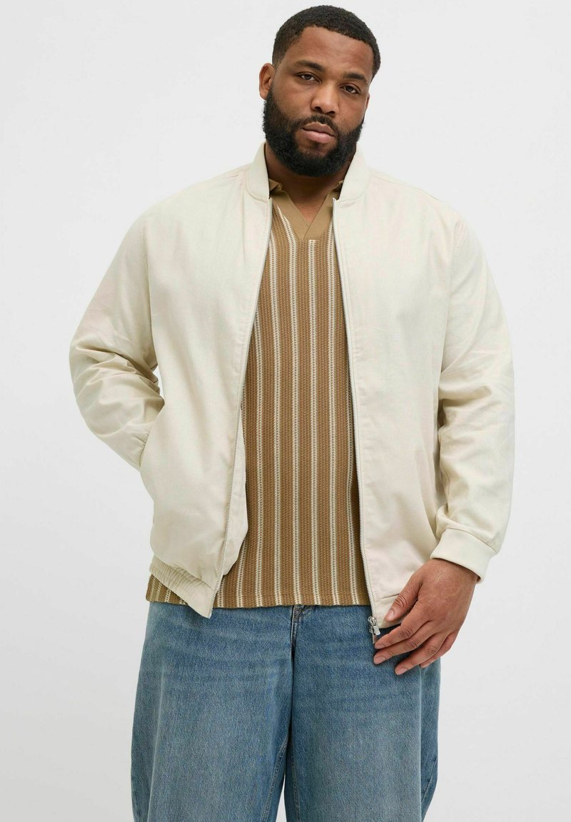 Cream bomber jacket with ribbed cuffs and collar, paired with a brown striped knit shirt and blue denim jeans.