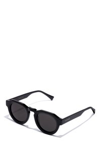 Schwarze Sonnenbrille mit runden Gläsern, dicken Rahmen und einem einzigartigen, kantigen Design. Verfügt über dezente Markenkennzeichnungen an der Seite und eine glatte Kunststoffoberfläche.