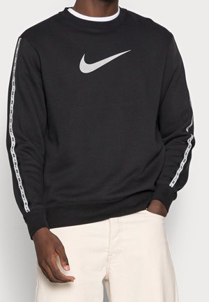 Mężczyzna ubrany w czarną bluzę Nike z białym logo na piersi i taśmą z logo na rękawach, w połączeniu z jasnymi spodniami.