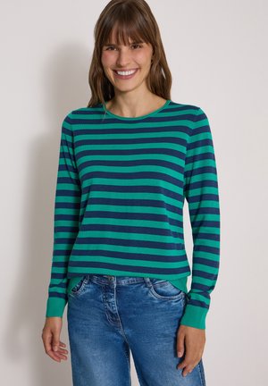 Kvinde smilende, iført en langærmet grøn og marineblå skjorte med horisontale striber og blå denimjeans, stående mod en ensfarvet baggrund.