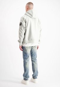 Lichtgrijze hoodie-jack met een gladde textuur, voorzien van een zwart embleem op de linkerarm. Draagt lichtblauwe distressed jeans en witte sneakers.