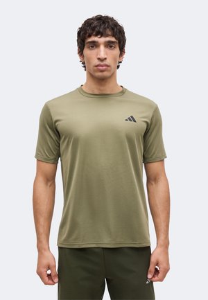 Man draagt een olijfgroen Adidas atletisch shirt met korte mouwen en bijpassende shorts, staand tegen een effen lichte achtergrond.