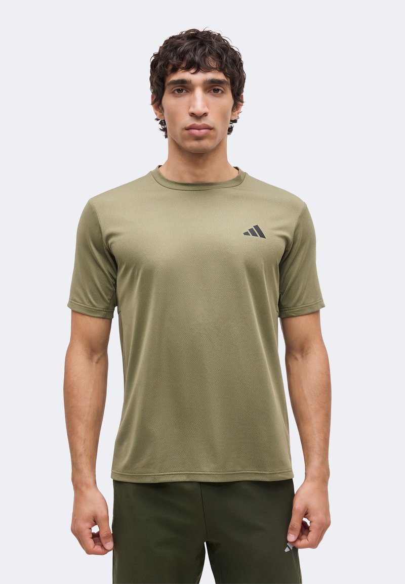 Homme portant un t-shirt de sport à manches courtes Adidas vert olive et un short assorti, debout devant un fond clair uni.