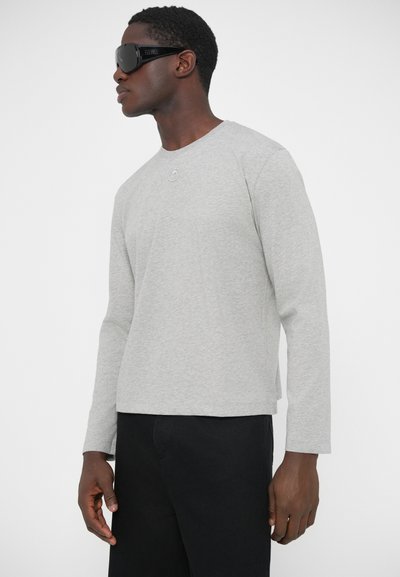 Sivá košeľa s dlhým rukávom a okrúhlym výstrihom, textúrovaná látka a jemný detail loga na hrudi, v kombinácii s čiernymi nohavicami a slnečnými okuliarmi.