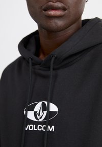 Sort hættetrøje lavet af blødt stof med et hvidt Volcom-logo. Leveres med snoretrækslukning og design med kengurulomme.