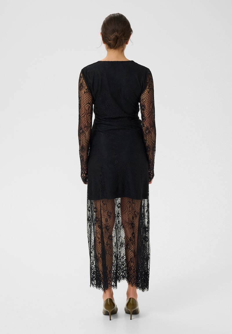Robe en dentelle noire à manches longues, avec une jupe transparente et des motifs floraux élaborés, se terminant sous le genou, associée à des talons vert olive.