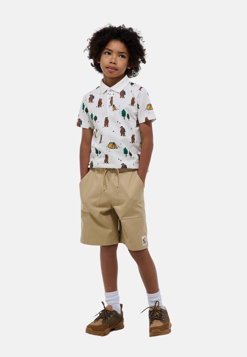 Polo blanc à manches courtes avec impression d'ours, d'arbre et de tente ; short beige avec poches ; chaussures marron ; chaussettes blanches.
