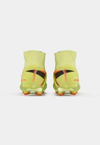 Botas de fútbol amarillas de caña alta con logotipos de Nike en negro y naranja, con tacos naranjas y suelas translúcidas, mostradas desde la parte trasera sobre un fondo blanco.