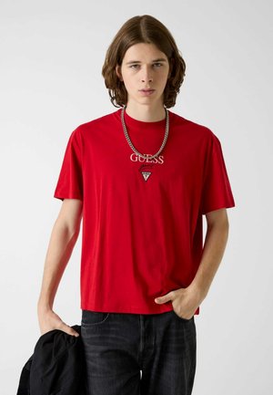 Joven con cabello castaño hasta los hombros lleva una camiseta roja de Guess, collar de cadena plateada, vaqueros negros, sosteniendo una chaqueta negra en una mano.