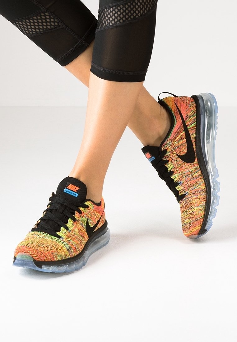 Jambes en leggings noirs en mesh, portant des baskets multicolores Nike Flyknit Max avec des semelles transparentes à coussin d’air, debout sur un sol clair.