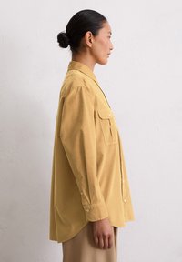 Femeie cu părul închis la culoare prins în coc, îmbrăcată cu o cămașă din bumbac galben muștar, lejeră, și pantaloni bej, stând de profil împotriva unui perete alb simplu.