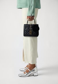 Borsa a tracolla nera trapuntata con lucchetto e un pompon soffice, abbinata a pantaloni bianchi aderenti e scarpe con tacco argentate.