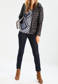 Veste matelassée noire, haut bleu et blanc à motifs, jean foncé et bottines marron. La veste est dotée d'une fermeture éclair et d'une capuche.