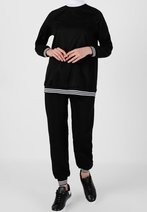 Osoba ubrana w czarną bluzę z długim rękawem i spodnie z białymi paskami na mankietach i dole, w czarnych trampkach na jednolitym szarym tle.