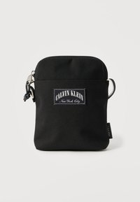 Μαύρη υφασμάτινη τσάντα με φερμουάρ, που διαθέτει λευκή ετικέτα με το λογότυπο Calvin Klein, λεία υφή και ρυθμιζόμενο λουρί στο πλάι.