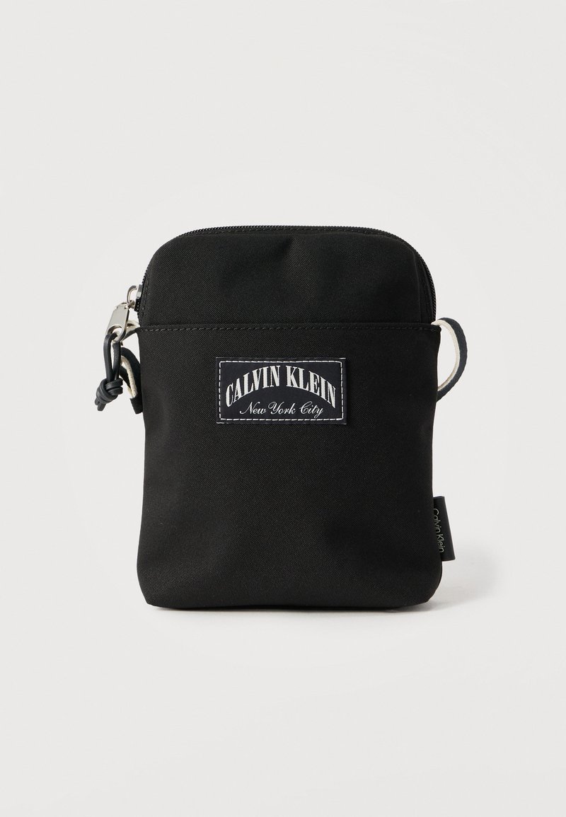 Μαύρη υφασμάτινη τσάντα με φερμουάρ, που διαθέτει λευκή ετικέτα με το λογότυπο Calvin Klein, λεία υφή και ρυθμιζόμενο λουρί στο πλάι.