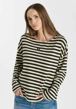 Donna con lunghi capelli castani che indossa un maglione a righe nere e crema e jeans blu, mani nelle tasche davanti, sfondo neutro.