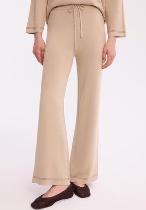 Beżowe dzianinowe spodnie o szerokich nogawkach z przewiązaniem w pasie, noszone z ciemnobrązowymi, plecionymi płaskimi butami.