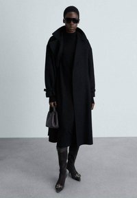 Cappotto di lana nero con ampi revers e polsini arrotolati, abbinato a un dolcevita nero, stivali al ginocchio e una piccola borsa scura.