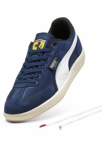 Marinblå ruskind- og stofsneaker med hvide detaljer, en struktureret cremefarvet gummisål og logodetaljer nær snørebåndene. Inkluderer snørebånd med røde spidser.