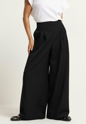 Frau trägt hoch taillierte, weit geschnittene schwarze Hose kombiniert mit weißem ärmellosem Oberteil und schwarzen spitz zulaufenden flachen Schuhen.