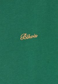 Grønt stoff med en teksturert overflate, med en brodert logo "Blkvis" i gull nederst til høyre. Enkel og ren design.