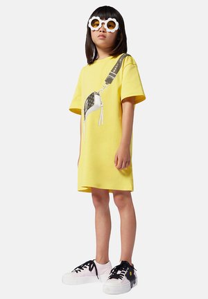 Robe t-shirt surdimensionnée jaune avec un graphique d'un sac à main, portée avec des baskets blanches et noires et des lunettes de soleil blanches en forme de fleur.