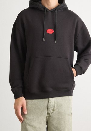 Osoba ubrana w czarną bluzę z kapturem z przednią kieszenią i czerwoną naszywką z logo Hugo, w połączeniu z jasnobeżowymi spodniami, stojąca na jednolitym tle.