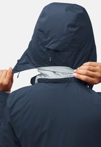 Veste coupe-vent marine avec capuche, comprenant un intérieur gris clair contrastant. Le col dispose d'une fermeture éclair et d'une fermeture Velcro pour l'ajustabilité.