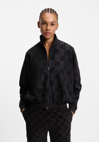 Sort lynlåstrøje og bukser med præget logomønster. Stoffet virker blødt med en tekstureret finish. Ribstrikkede ærmekanter og talje.