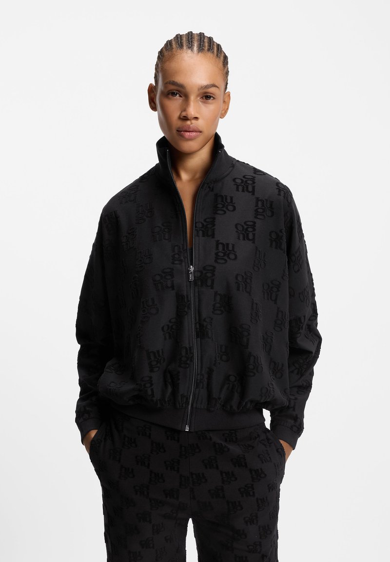 Sort lynlåstrøje og bukser med præget logomønster. Stoffet virker blødt med en tekstureret finish. Ribstrikkede ærmekanter og talje.