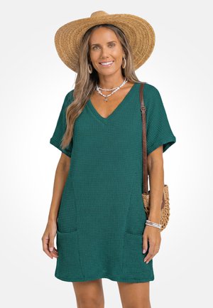 Vrouw draagt een groene, gestructureerde korte mouwen jurk, grote strohoed, schelpenketting, creooloorbellen, armbanden en een geweven schoudertas.